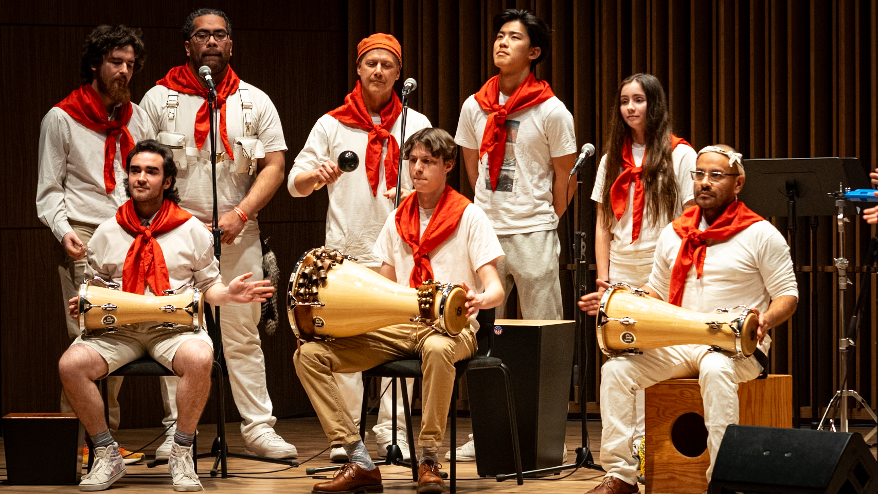 Ensemble of musicians in white with red scarves playing hand drums on stage.