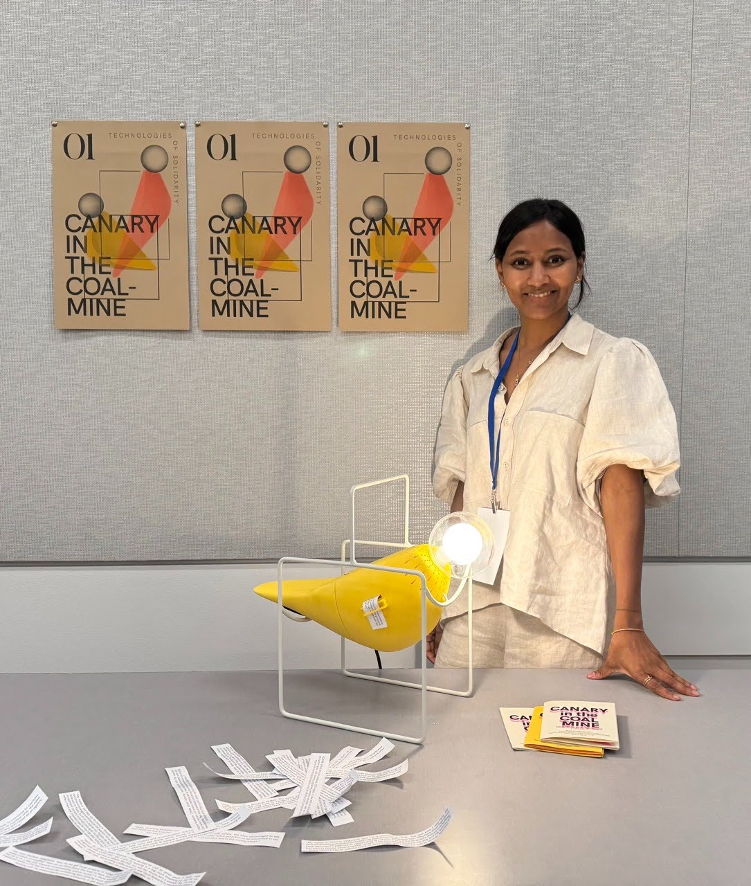 Smiling woman in studio with a yellow paper model, scattered paper strips, and posters