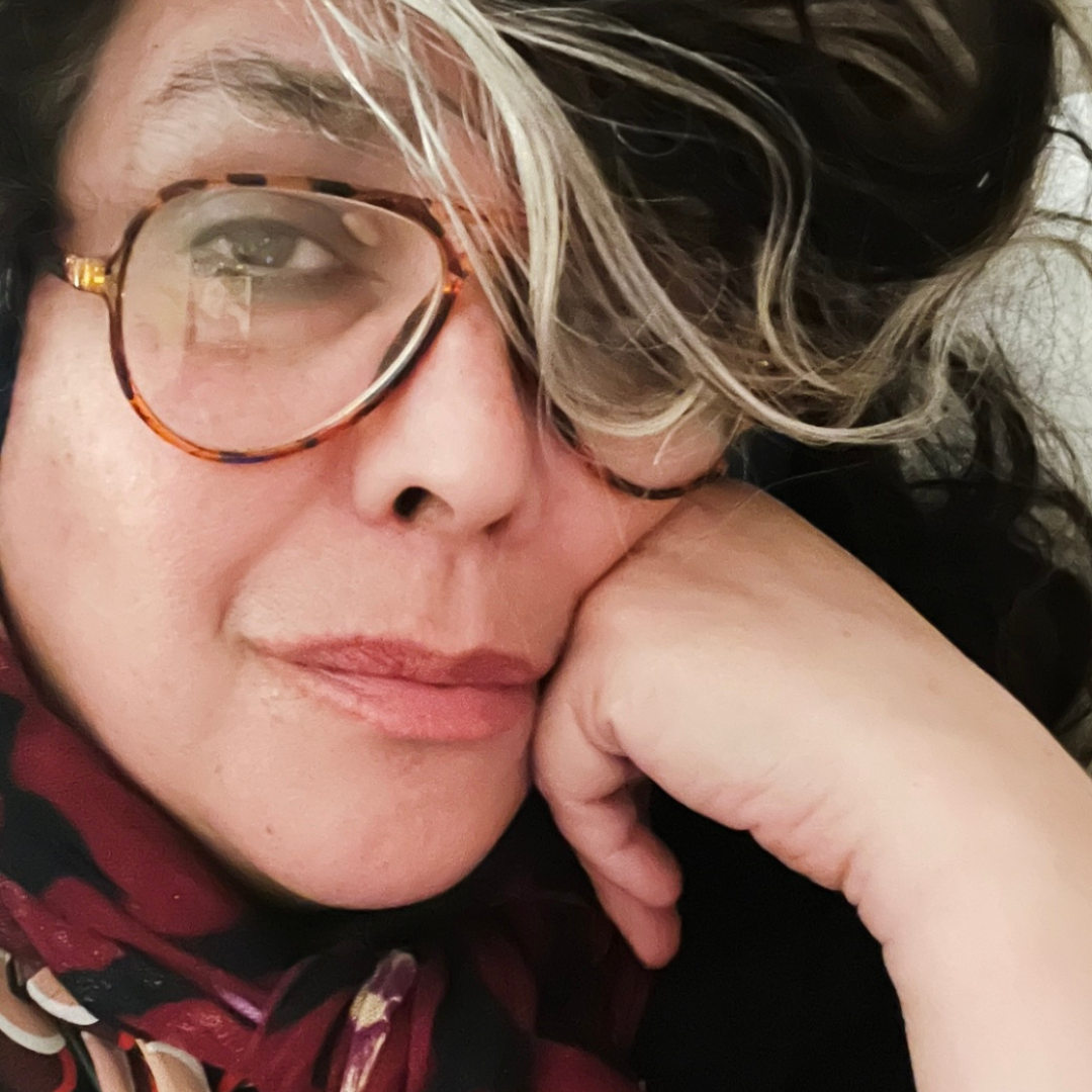 Close-up photo of person with tortoiseshell glasses, silver-streaked hair, hand on cheek.