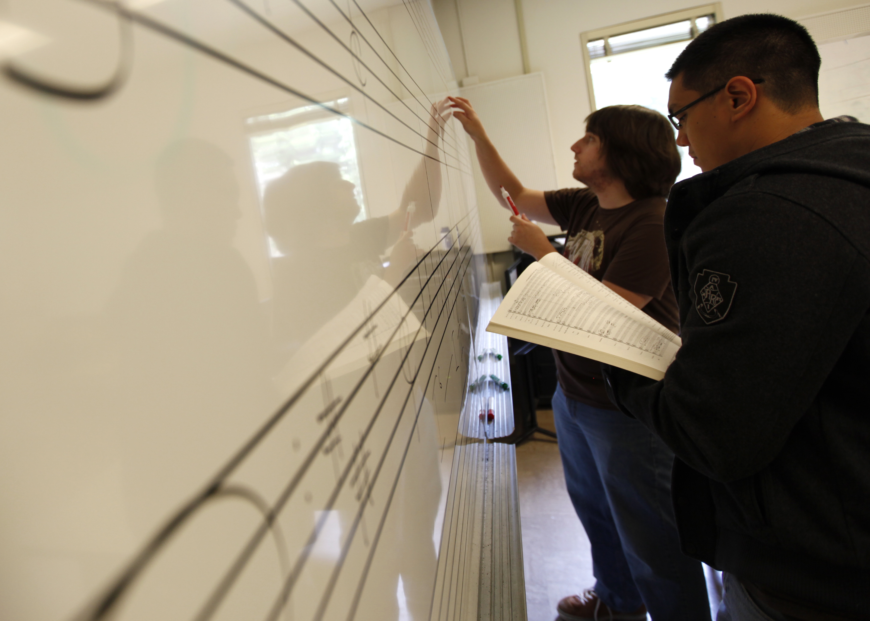 Two students writing musical staff lines on a classroom whiteboard, one holding a full score