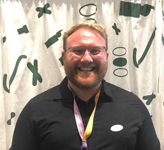 Smiling bearded man with glasses wearing black shirt and lanyard, green abstract backdrop