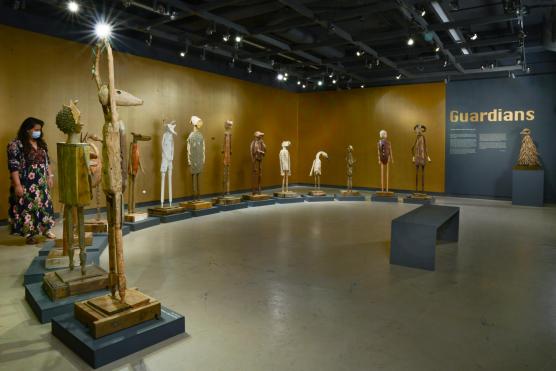 Wooden guardian sculptures on stands along a curved gallery wall; visitor nearby.