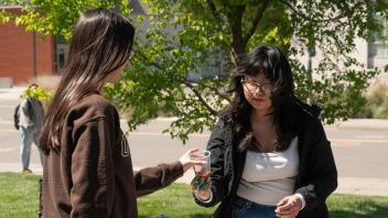 Two women exchange art supplies at an outdoor table on a sunny campus