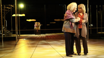 Two women embracing on a warmly lit theater stage with reflective floor