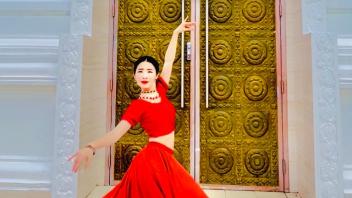 Female dancer in flowing red gown posing gracefully before ornate golden doors
