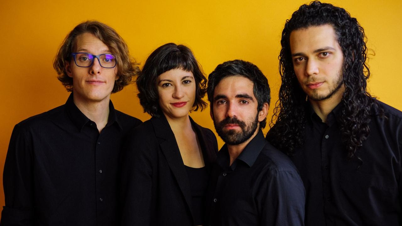 Studio portrait of four people in black clothing against a yellow background, neutral expressions