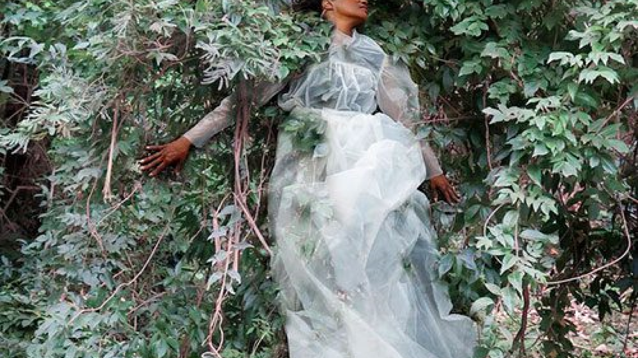 A performer draped in a flowing pale garment stands amid dense green foliage, photographed in a soft, blurred artistic style.