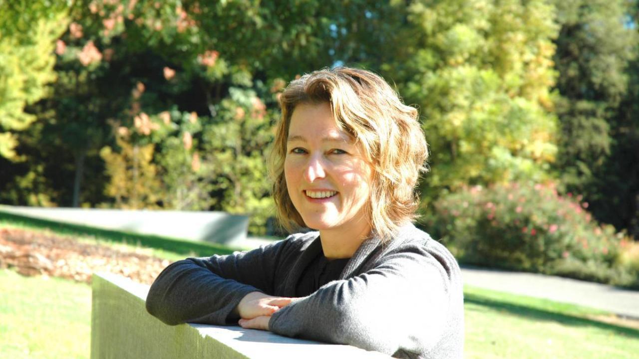 Smiling woman with curly blonde hair, leaning on a railing outdoors with trees in the background.