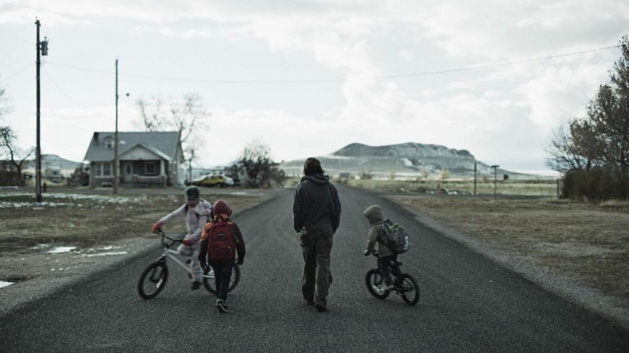 Film still from "The Killing of Two Lovers" showing a man and two children walking down a rural road under an overcast sky, with a small town visible in the distance.
