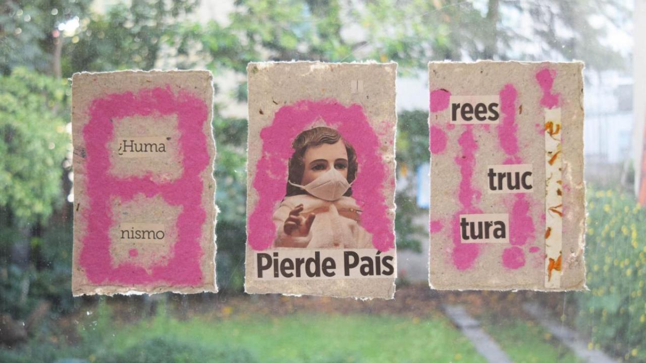 Three pink paper banners strung on a line outdoors, each hand-lettered with text and small painted figures, set against blurred green foliage.