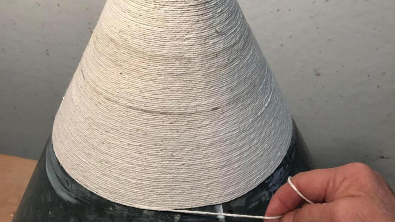 White conical woven sculpture on a black base, with a hand delicately touching its rim edge.