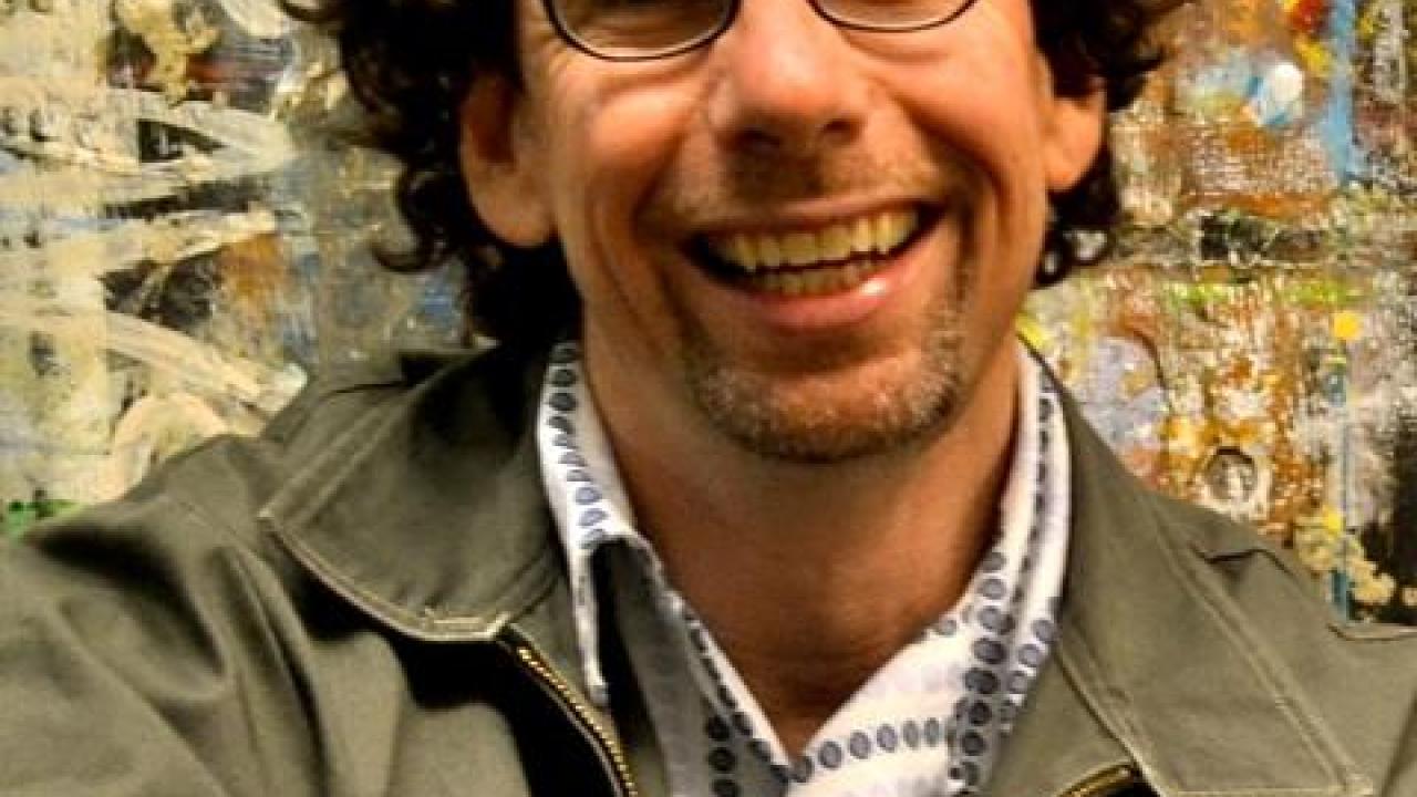 Smiling man with curly dark hair and glasses, wearing a light-colored jacket outdoors.