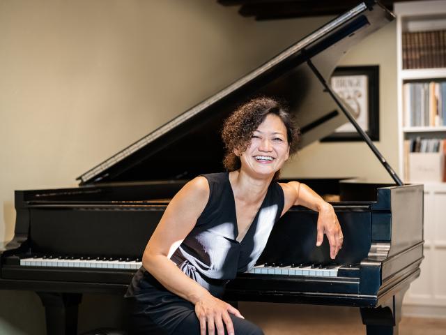 Smiling woman with short curly hair in black-and-white dress leaning on a grand piano in cozy room