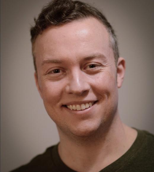 Smiling young man in gray shirt, outdoor headshot with blurred green background.