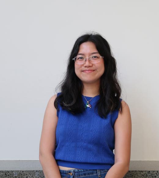 Young woman with glasses in a blue sleeveless top, seated and smiling against a white wall