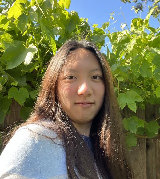 Young person with long dark hair in blue top standing by green vines and bright blue sky