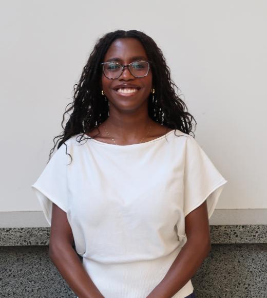 Smiling woman with long dark braids, glasses and white blouse seated against a pale wall
