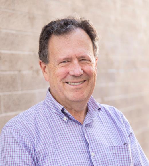 Headshot of smiling middle-aged man in purple checkered shirt by tan brick wall