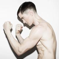 Profile view of Cassils' body, fists closed, and arms in an upright wrestling-like position against a neutral white background.