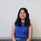 Young woman with glasses in a blue sleeveless top, seated and smiling against a white wall
