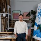 Artist in cream sweater standing in cluttered studio between large canvases