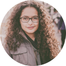 Young woman with brown curly hair and glasses, slight smile, outdoor city background (photo)