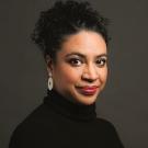 A woman with curly hair, black turtleneck, white earrings, dark background.