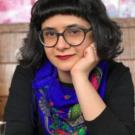 Portrait of Aida Shirazi wearing glasses and a bold blue scarf, against a soft floral background.