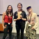 Three women standing together indoors, smiling, one holding a small trophy, with a projector screen in the background.