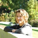 Laurie San Martin leaning on a white railing outdoors, with sunlit trees in the background.