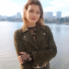 Young woman in olive jacket and black lace top, arms crossed, smiling by lakeside skyline