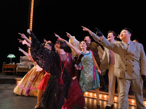 Actors in colorful costumes point upwards toward the audience.