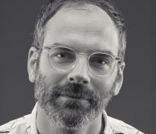Josh Faught (courtesy) A grayscale portrait photo of Josh Faught. He's wearing clear-rimmed eyeglasses and wearing a collared shirt with large thick-lined shapes on it.