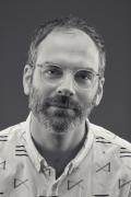 A grayscale portrait photo of Josh Faught. He's wearing clear-rimmed eyeglasses and wearing a collared shirt with large thick-lined shapes on it.
