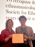 Two women standing together in front of a conference banner, one presenting an orange award certificate to the other.
